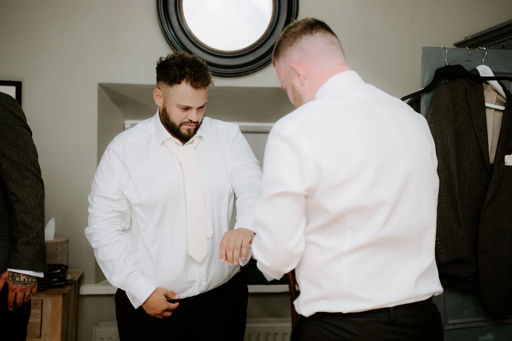 Two men in white shirts stand in a room with a round mirror on the wall as one of them adjusts the other's cuff