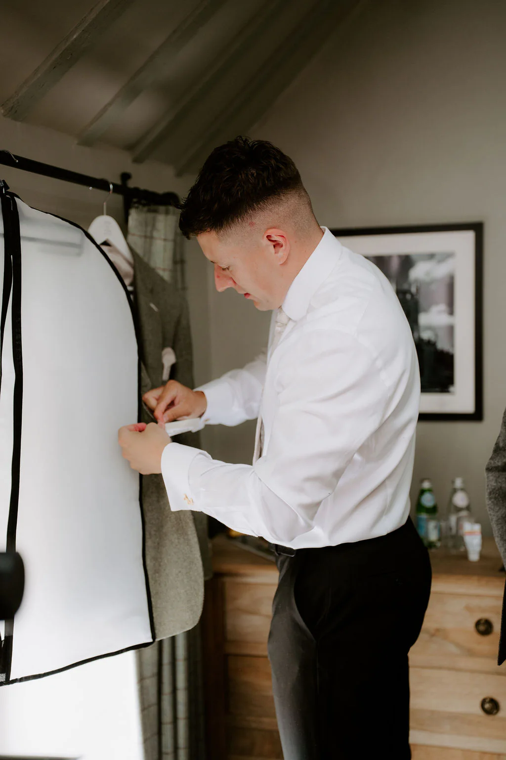 A man in a white shirt and dark trousers fastens a button beside a suit hanging in a garment bag in a pale room