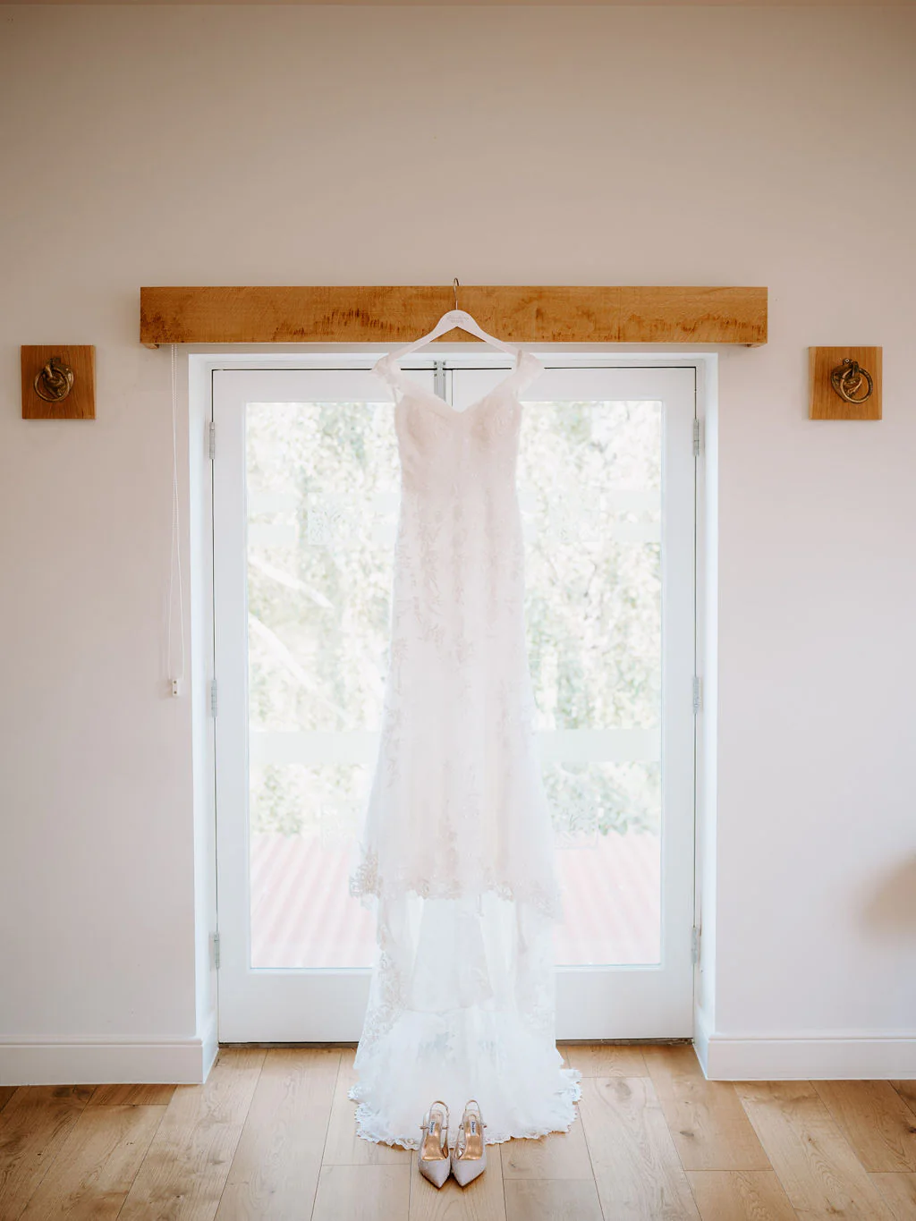 A lace wedding dress hangs from a wooden beam in front of a pair of glazed doors in a bright room