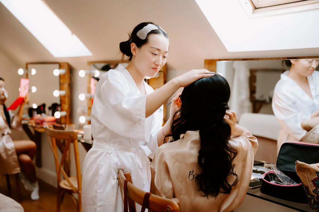 A woman in a white robe fixes the hair of a seated woman in a bright room with a bulb-lit mirror and several people beyond