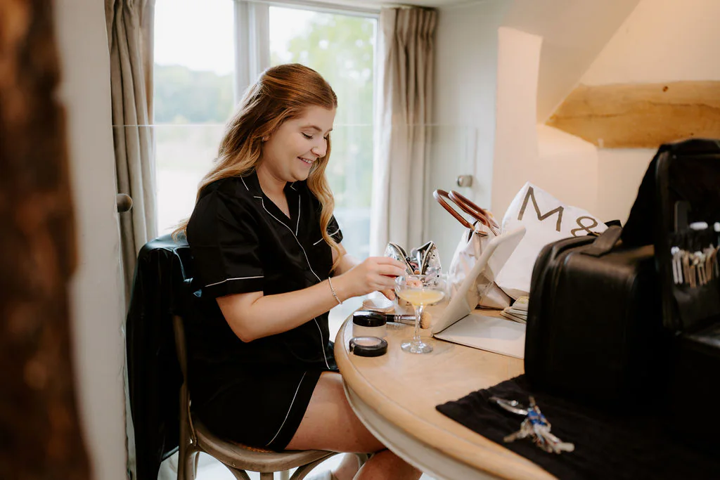 A woman in black pyjamas sits at a round table by a window with a glass of champagne and a garment bag beside her