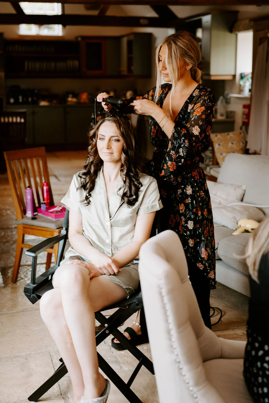 A bride in satin pyjamas sits on a director's chair in a bright farmhouse-style room while a woman in a floral robe pins up her hair
