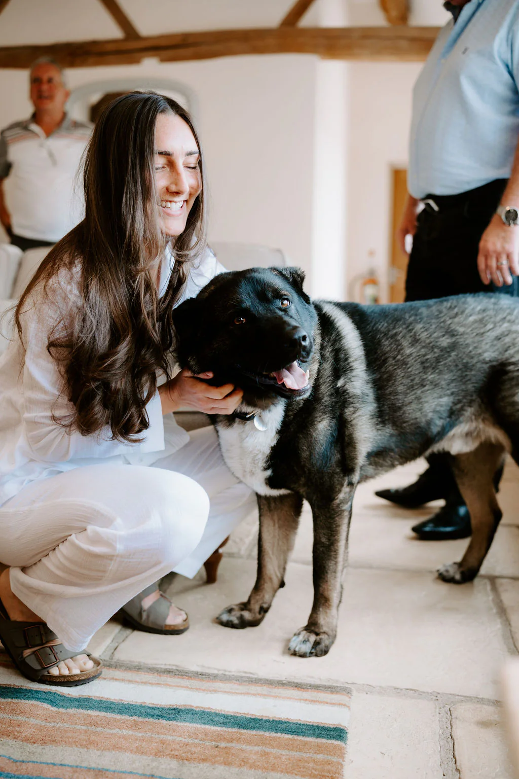 A smiling woman in white crouches to stroke a large dark dog on a stone floor with a striped rug