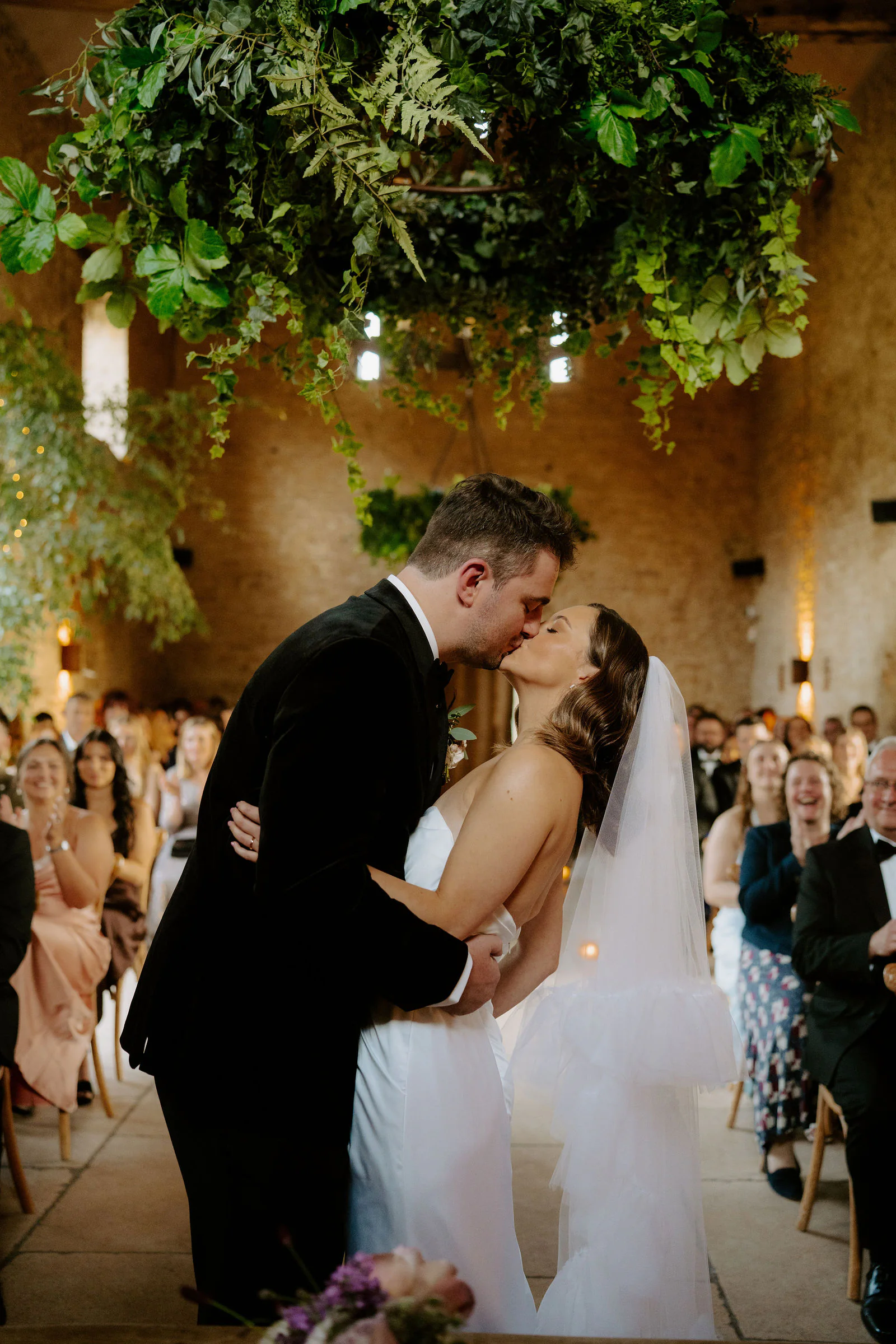 Lauren and Henry kissing at the end of their ceremony beneath a hanging garland of greenery inside a stone barn at Old Gore Barn near Cirencester, guests smiling in the rows behind them