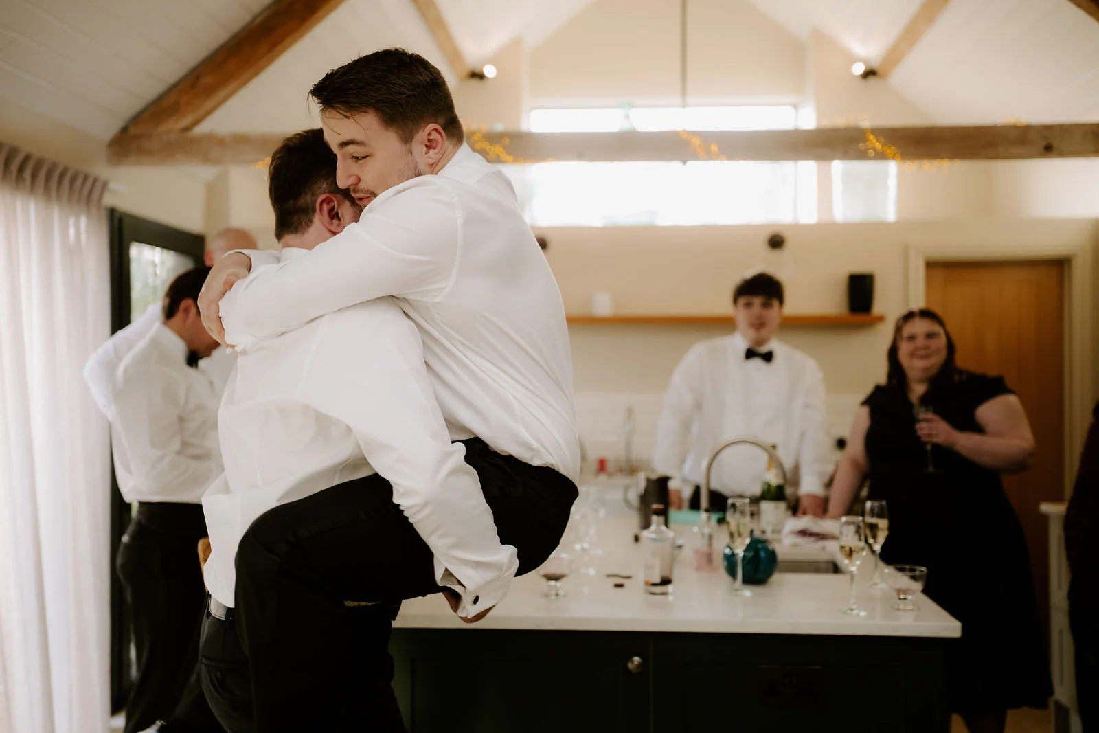 A man in white shirtsleeves lifts another man in a bear hug beside a kitchen island, with a man in a bow tie and a woman in a dark dress watching behind