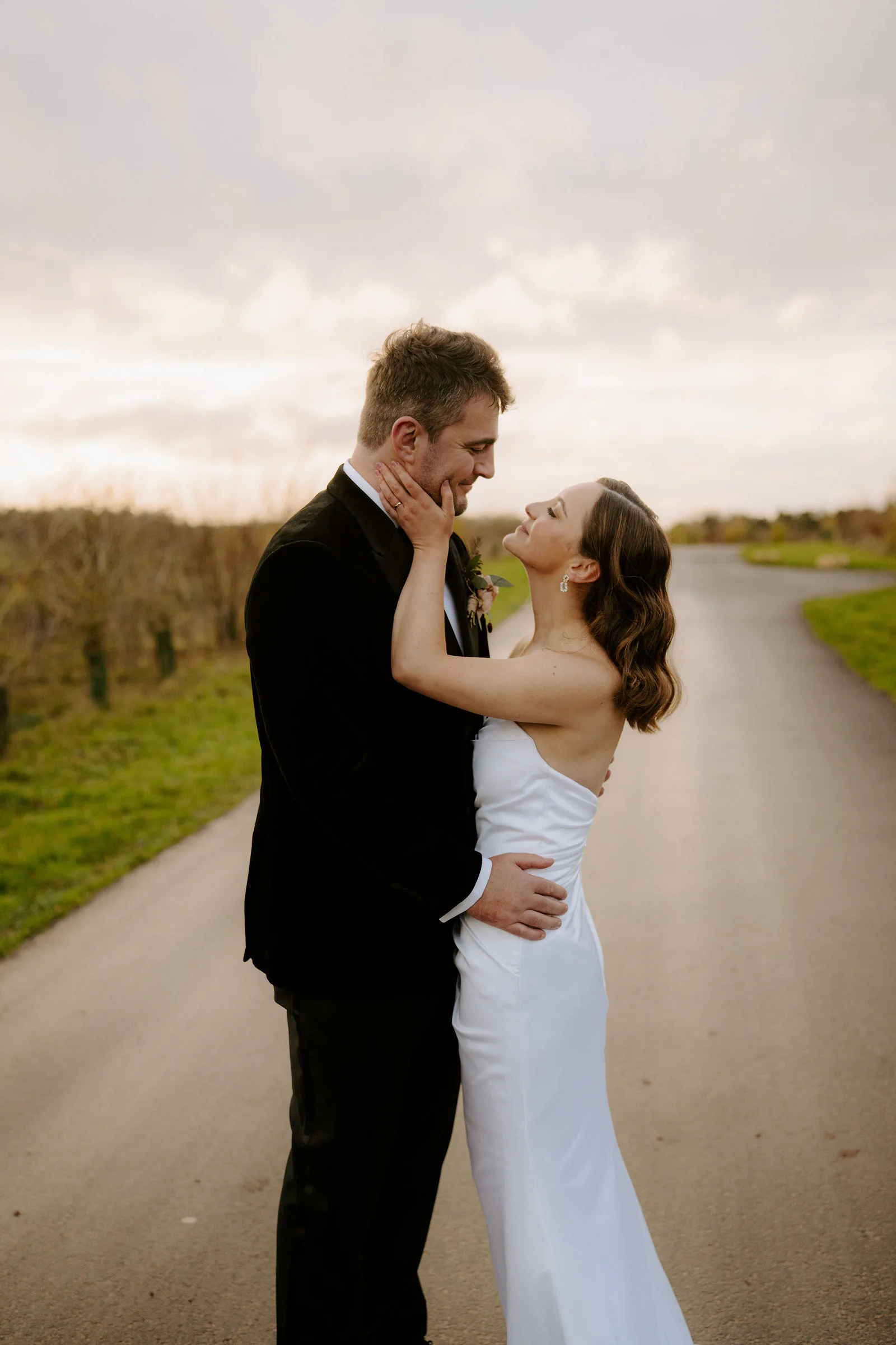 Henry holding Lauren's face as she looks up at him on a country lane beneath a pale evening sky near Old Gore Barn
