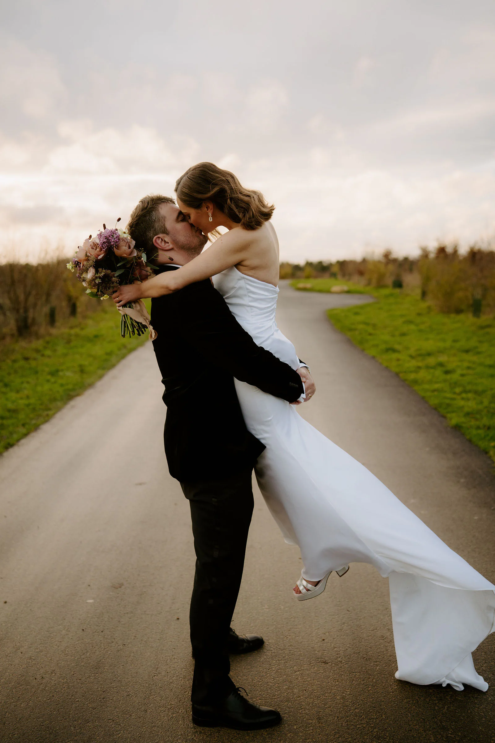 Henry lifting Lauren into a kiss on a country lane at dusk, her dress flowing, with grass verges and fields behind them near Old Gore Barn