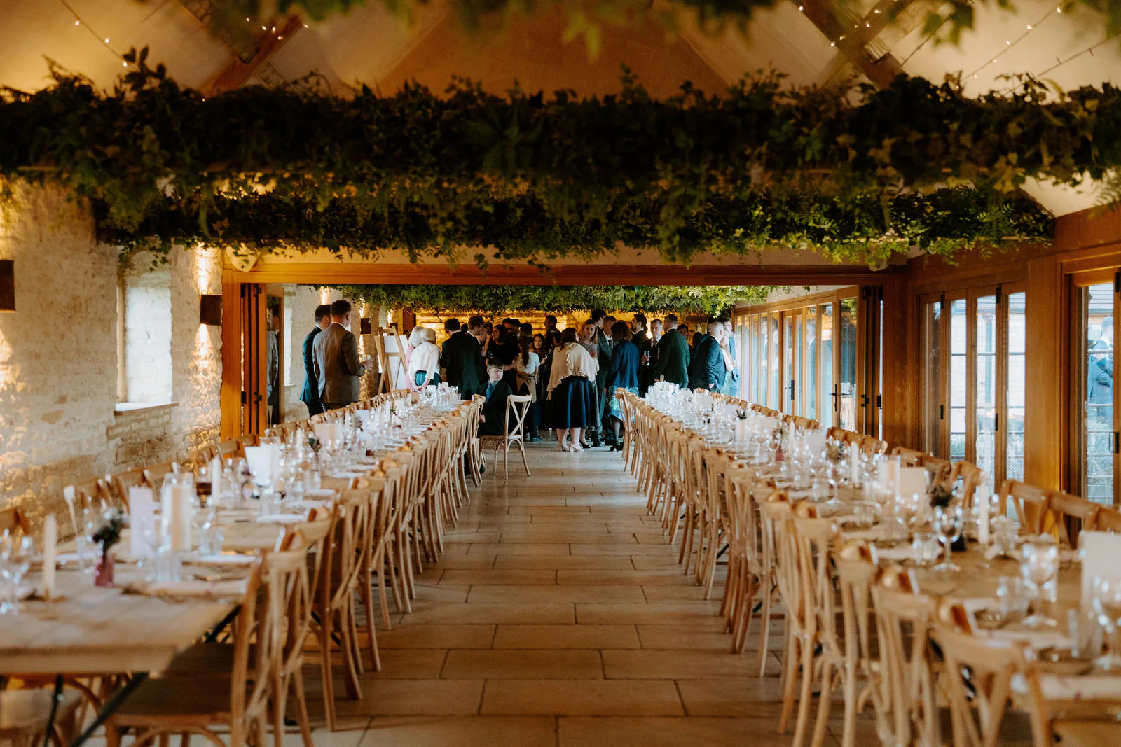 Long wooden tables laid with glasses and candles stretch down a stone barn beneath a ceiling of hanging greenery and lights at Old Gore Barn