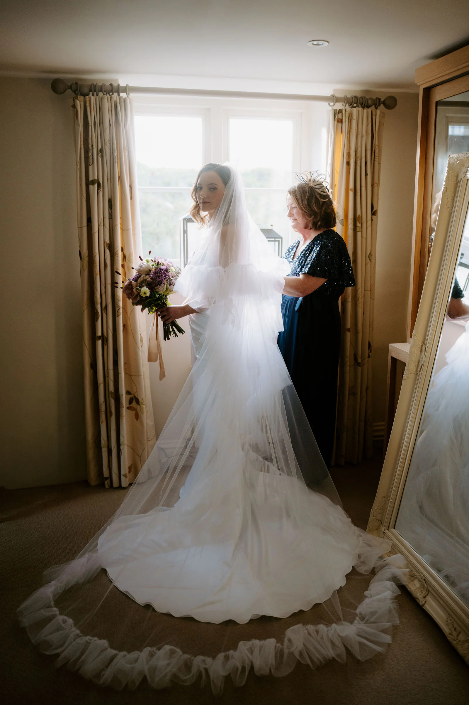 Lauren in a long veil holding a bouquet beside a window, a relative in a dark dress next to her, before the ceremony at Old Gore Barn