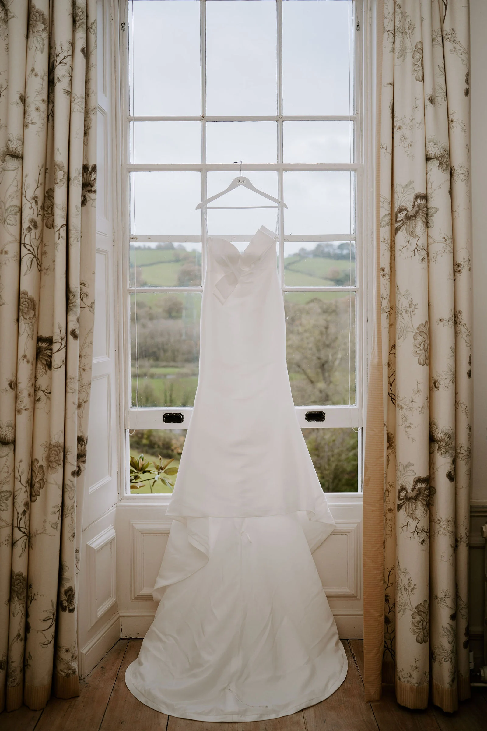 Wedding dress hanging gracefully in front of a grand Georgian sash window overlooking the Devon hills at Pynes House