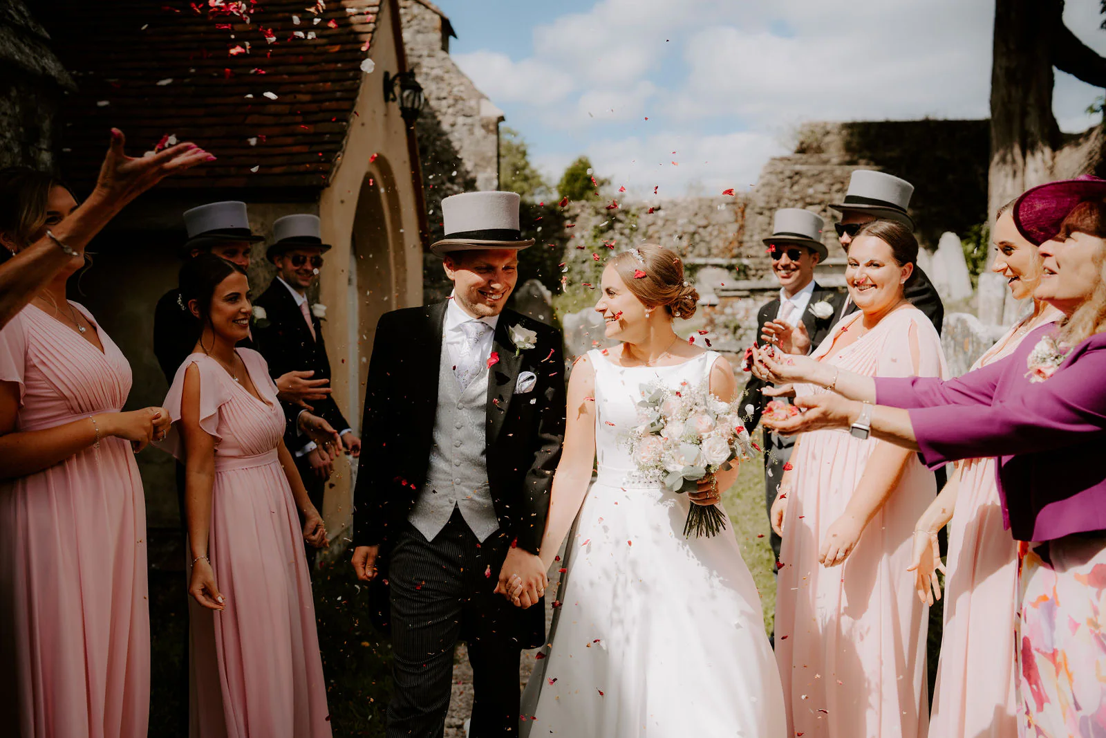 Chloe and Matt walk hand in hand through confetti outside a stone church, with bridesmaids in pink on one side and guests in top hats on the other