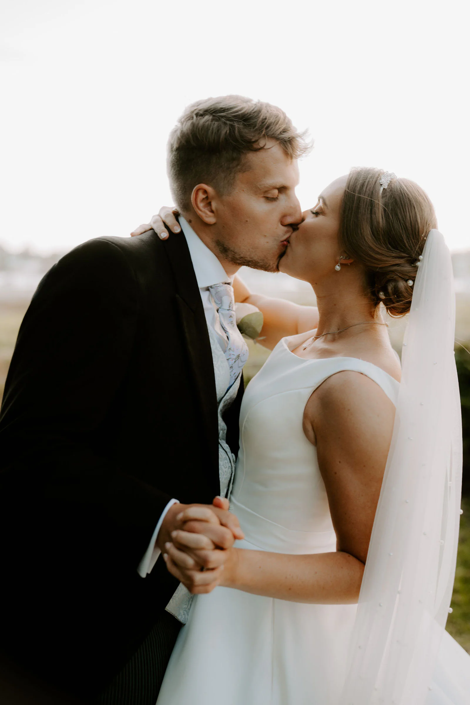Matt and Chloe kiss close together in warm evening light, her hand clasped in his, with water and moored boats softly blurred behind them