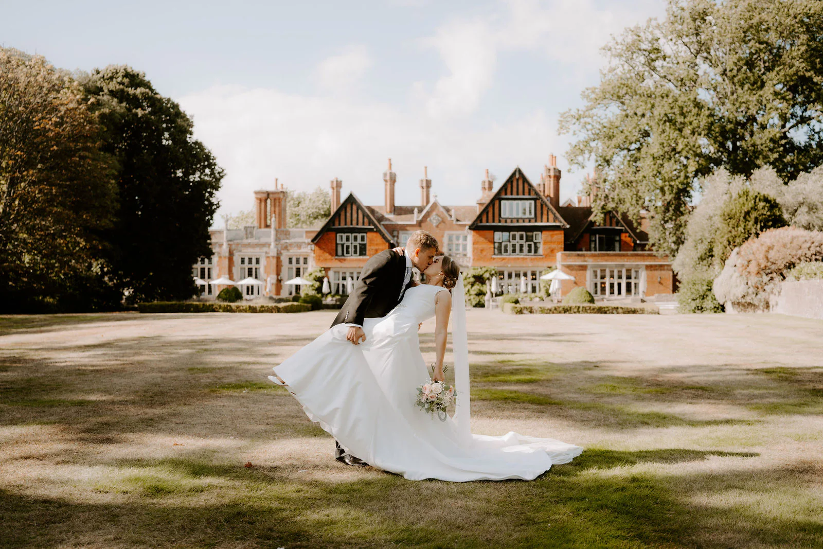Matt dips Chloe into a kiss on a wide lawn in front of a red-brick country house with tall chimneys