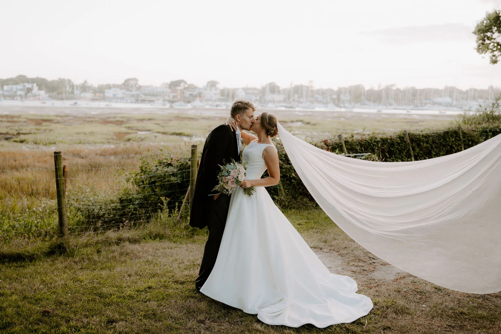 Chloe and Matt kiss on a grassy bank as her long white veil streams out in the wind, with an estuary and moored boats behind them