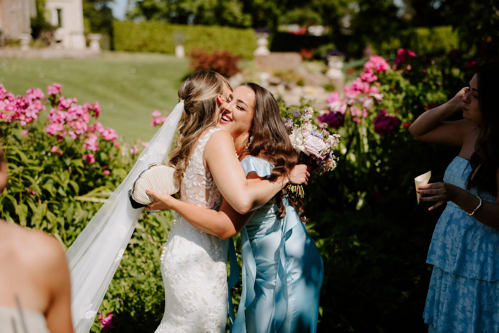 Faye in her veil hugs a bridesmaid in a blue dress among pink peonies in the garden at Parley Manor