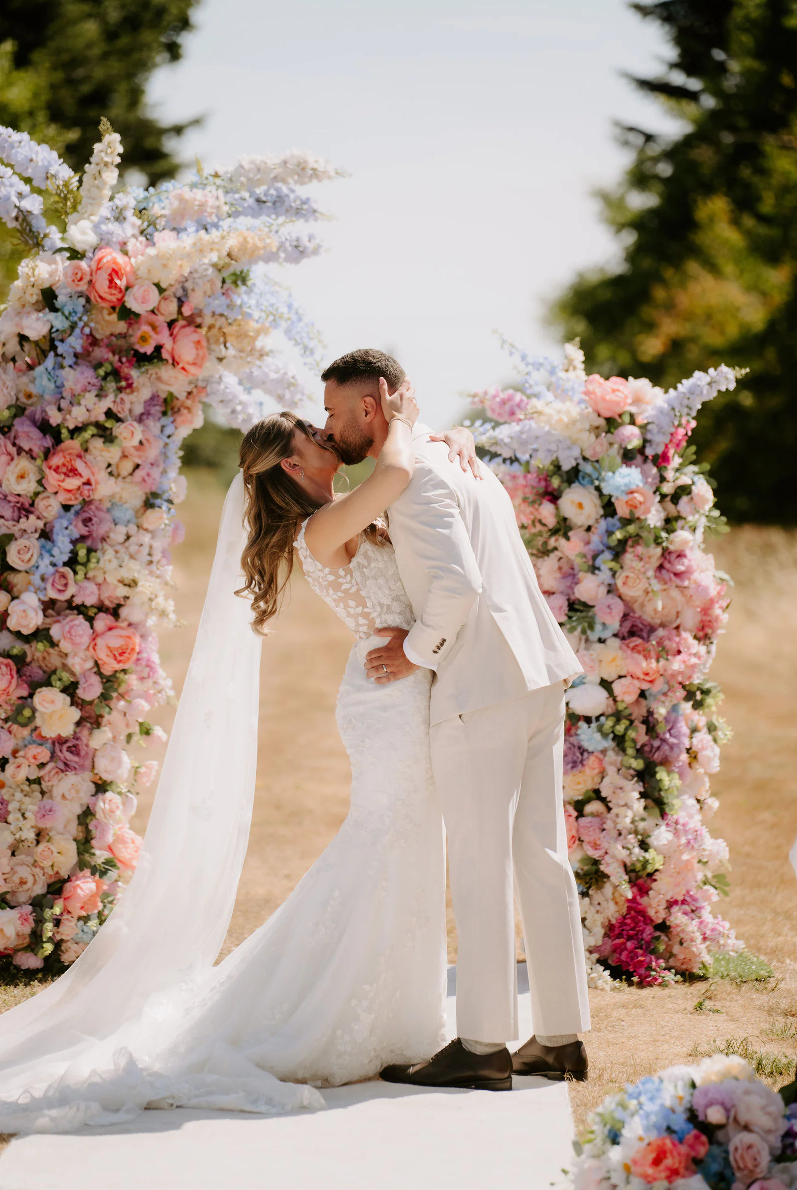 Faye and Sam kiss in front of a tall arch of pastel flowers on a white aisle runner at Parley Manor