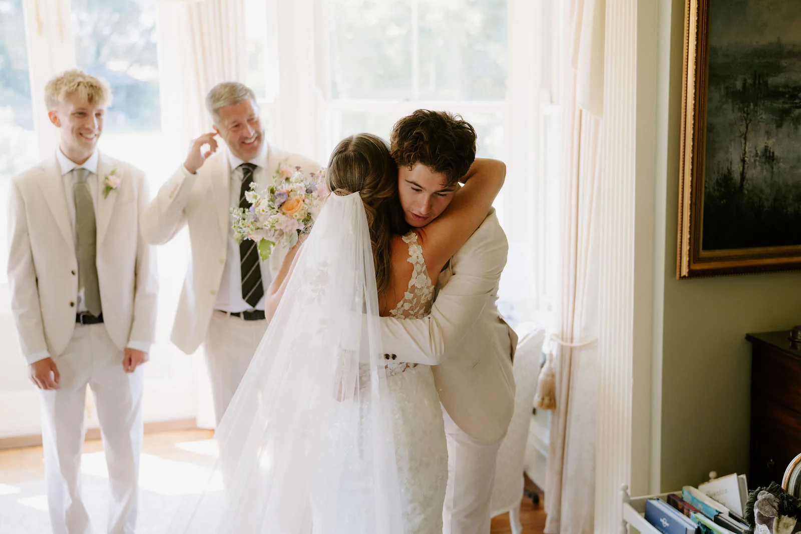 Faye in her veil hugs a man in a cream suit in a sunlit room while two other men in cream suits look on