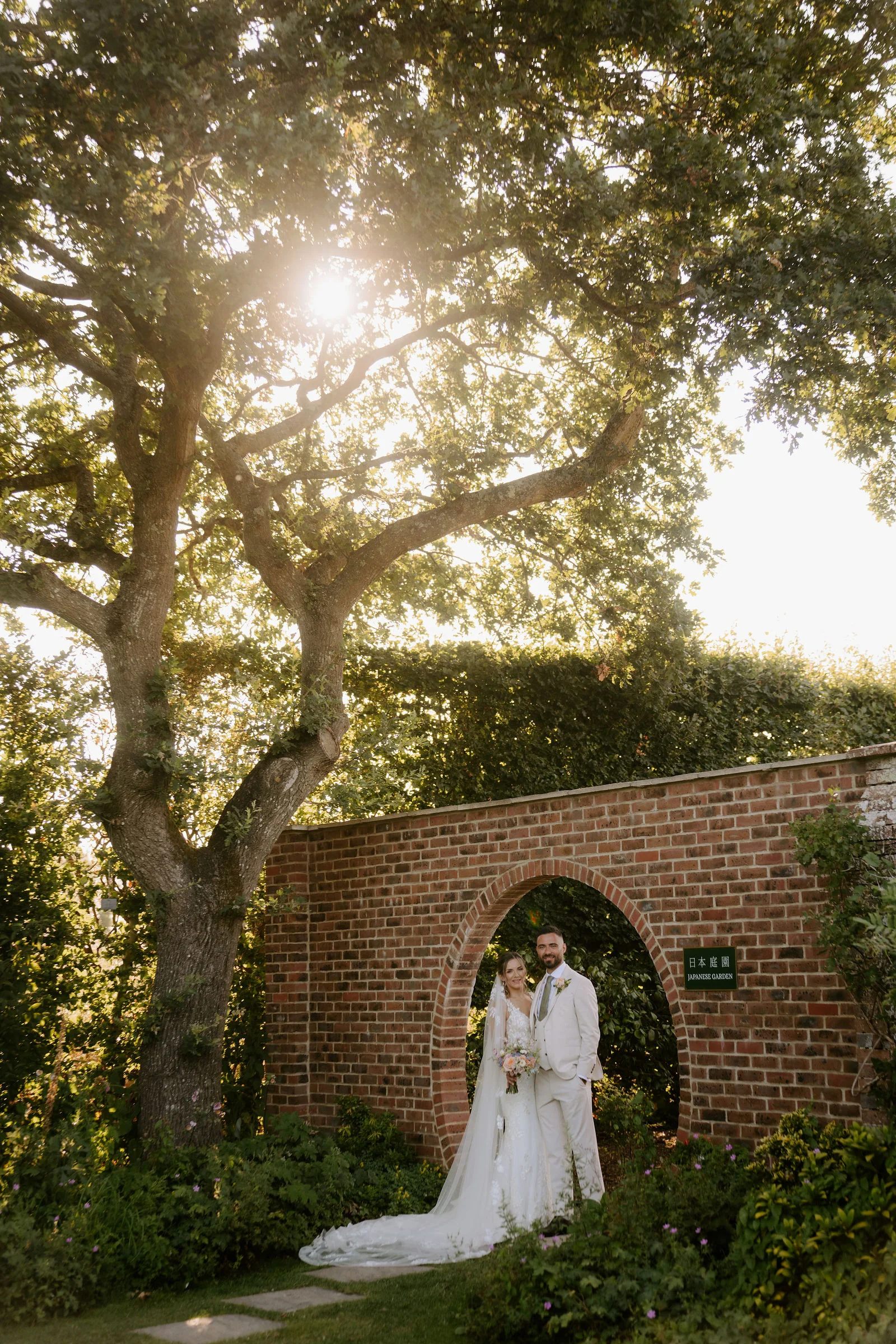 Faye and Sam beneath a circular brick doorway with sunlight through the trees at Parley Manor