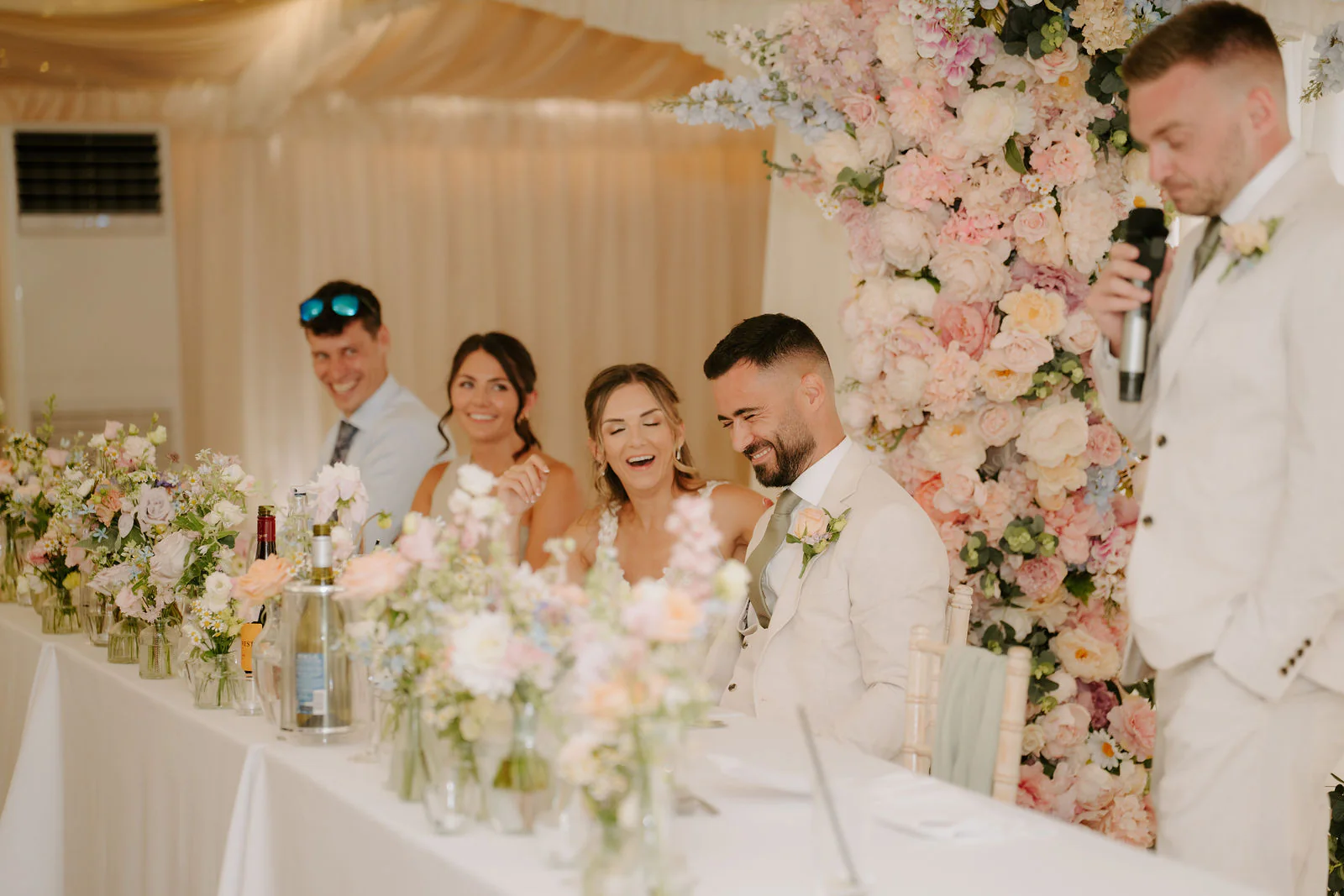 A groomsman gives a speech with a microphone while the wedding party laughs at the top table beside a wall of pastel flowers in the marquee at Parley Manor