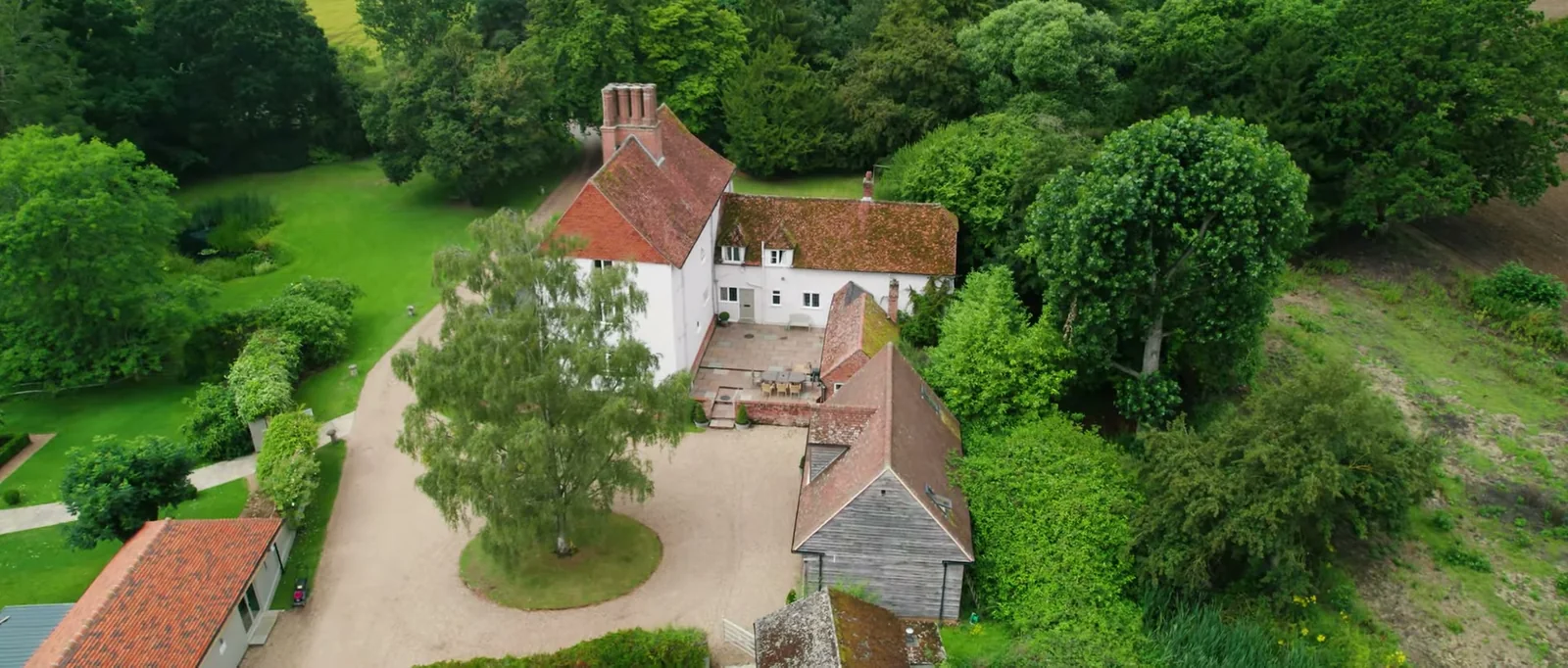An aerial view of a white timber-framed farmhouse with a red-tiled roof, a gravel drive and barns among trees and lawns at Houchins