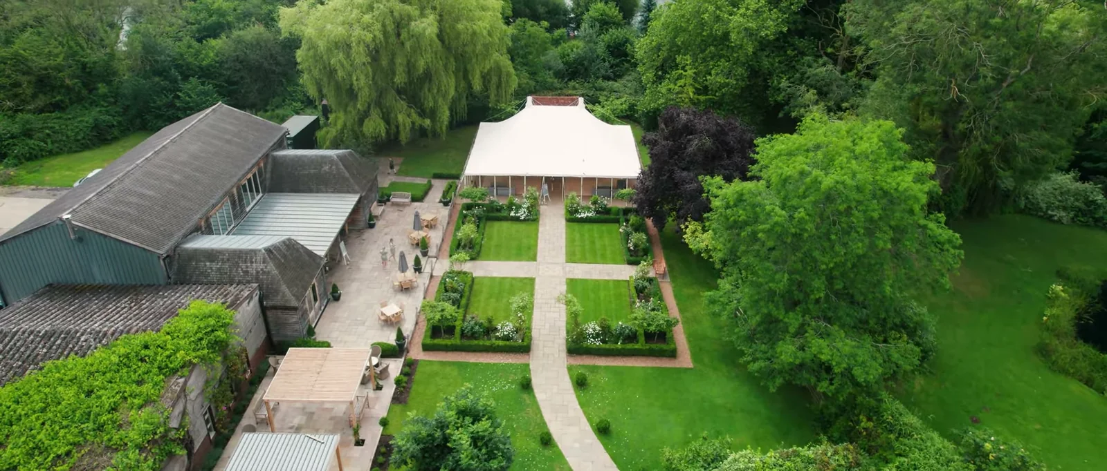 An aerial view of a long barn roof, a white marquee and a formal garden with grass paths and clipped beds at Houchins