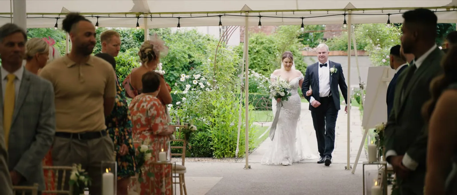 A bride in a lace dress carrying a bouquet walks down an aisle with a man in black tie under a marquee roof hung with festoon lights while guests stand either side