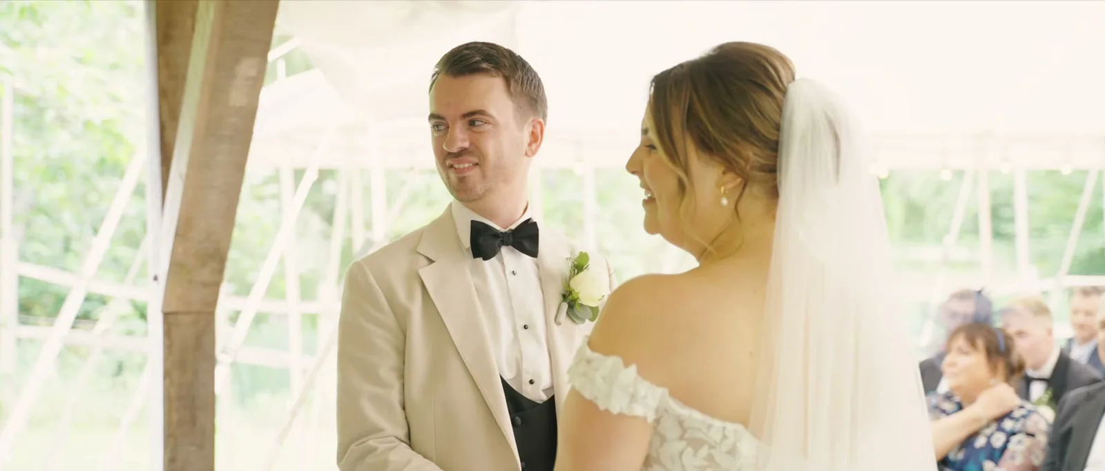 A groom in a cream jacket and black bow tie smiles at his bride in a veil during the ceremony under the marquee