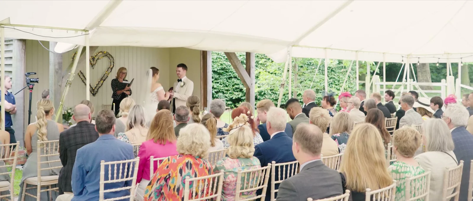 Guests sit on white chairs facing a celebrant and the couple under a white marquee, with a heart made of twigs on the wall behind them