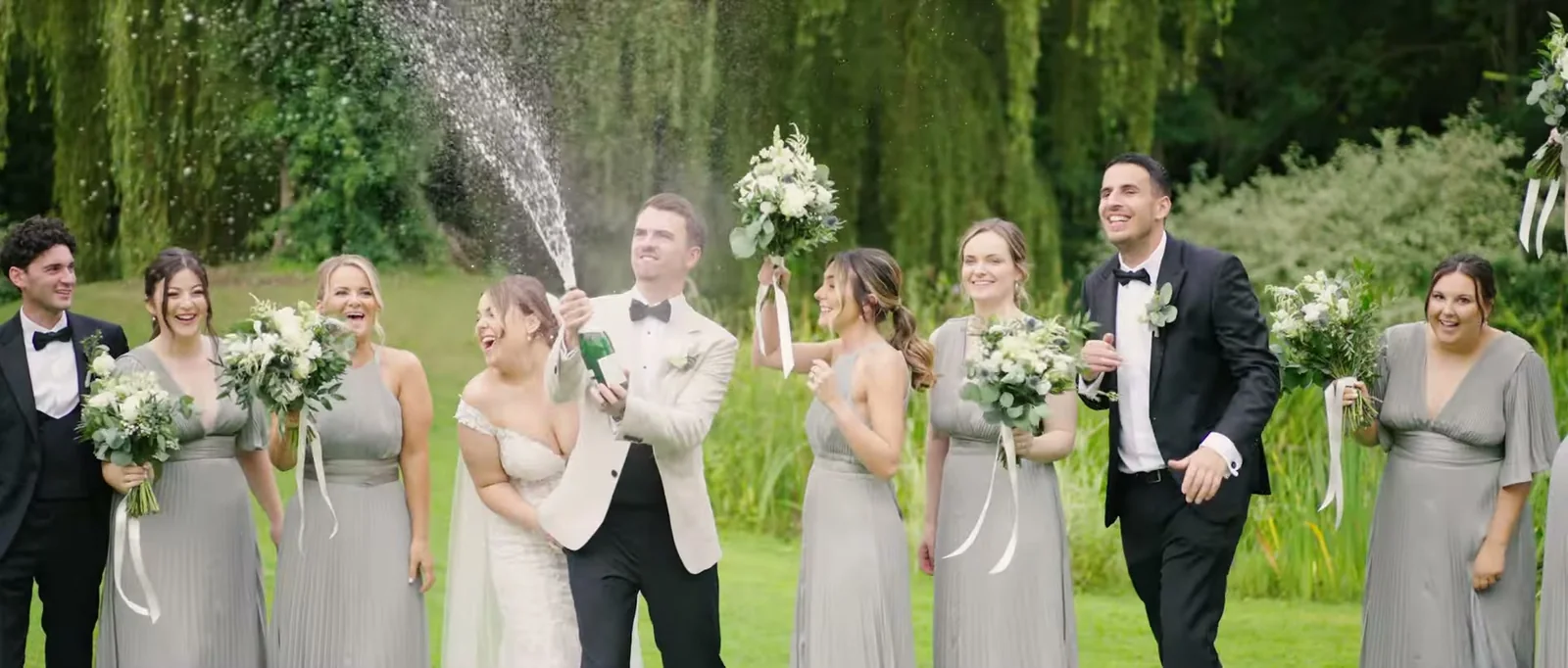 A groom in a cream jacket sprays champagne beside bridesmaids in grey and groomsmen in black on a lawn with a willow behind them