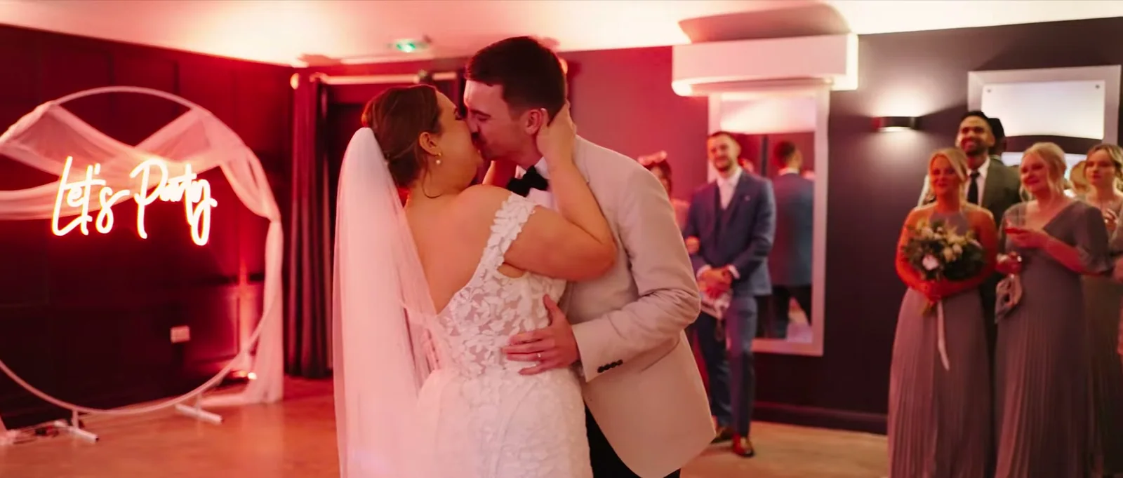 A couple kiss during their first dance in a dim room lit red, beside a neon sign that reads Let's Party, with bridesmaids watching