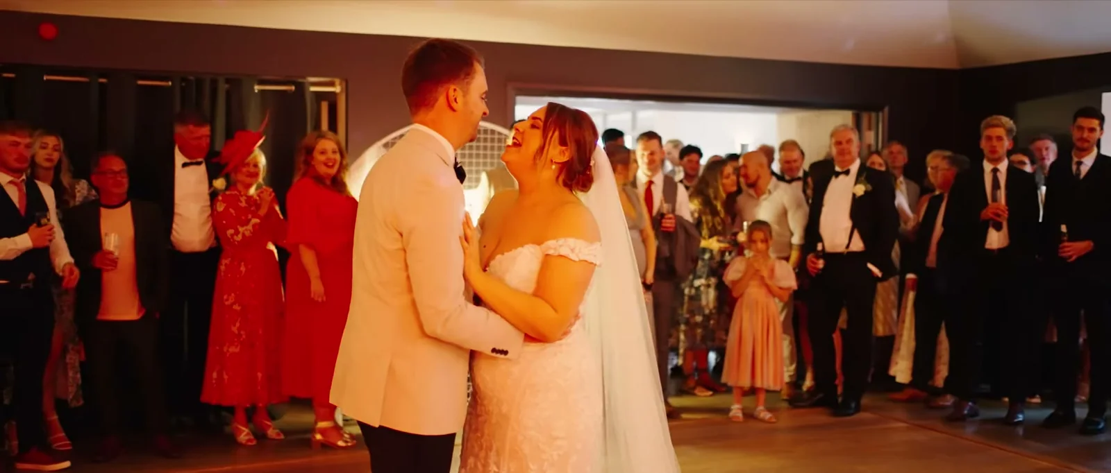 A groom in a cream jacket and a bride in a veil share their first dance while guests stand around them watching