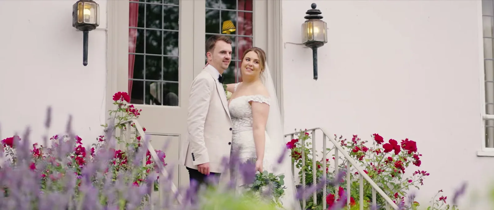 A groom in a cream jacket and a bride in a veil stand together against the white front of a farmhouse with two lanterns, lavender and red roses in the foreground at Houchins