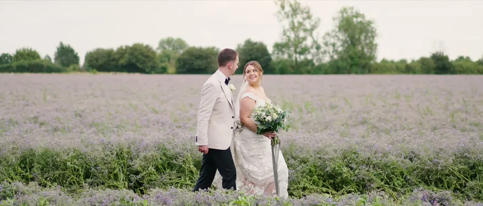 A groom in a cream jacket and a bride holding a bouquet walk through a field of pale purple flowers under a grey sky