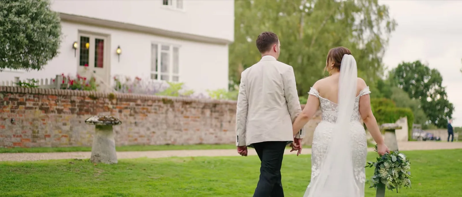 A couple walk away hand in hand across a lawn past an old brick wall, with the white farmhouse behind them