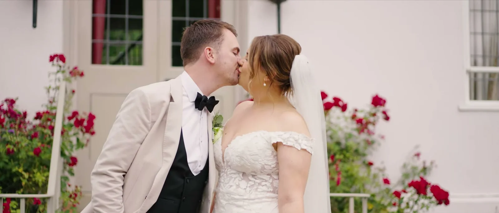 A groom kisses his bride beside a white door framed by red climbing roses
