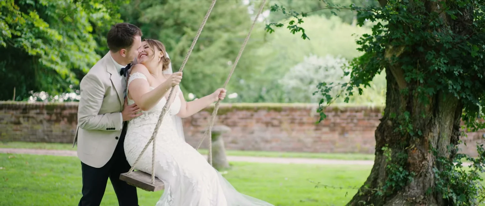 A groom in a cream jacket leans in to kiss his bride as she sits on a rope swing hung from a large tree, with a brick wall behind them