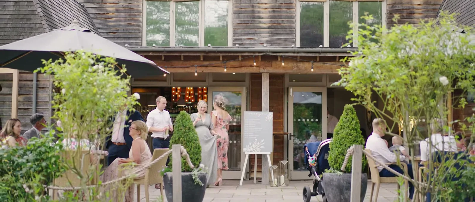 Guests gather on a terrace in front of a timber barn with parasols, string lights and planted pots