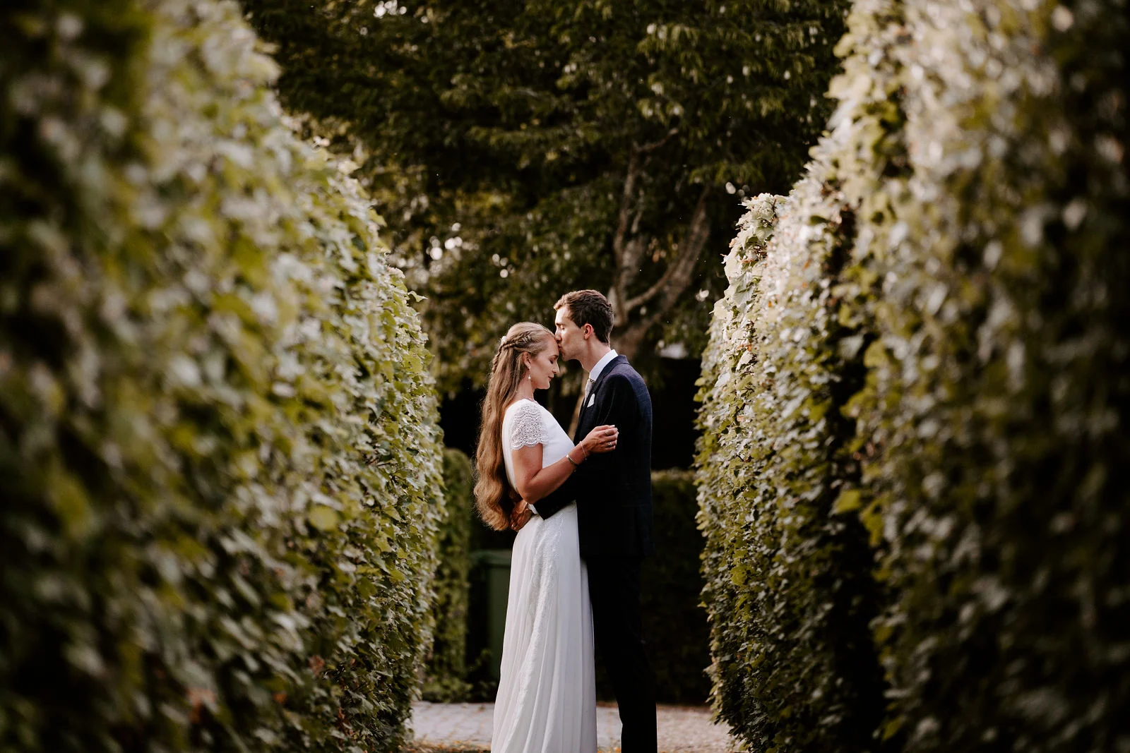 Tom and Sarah embracing between tall green hedges at Bury Court