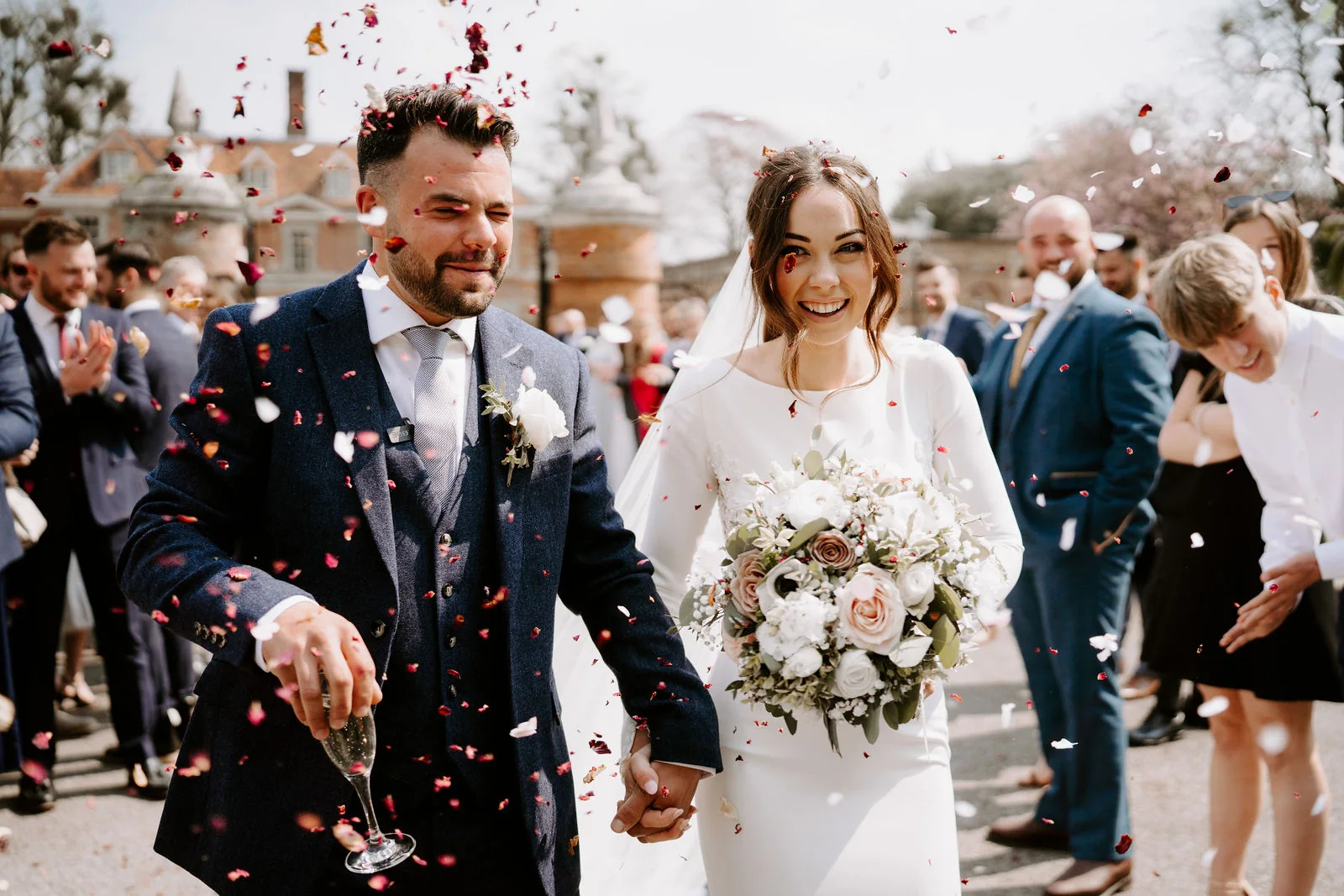 A groom in a navy suit and a bride with a bouquet of white and pink flowers walk through a shower of confetti as guests cheer behind them at Lainston House