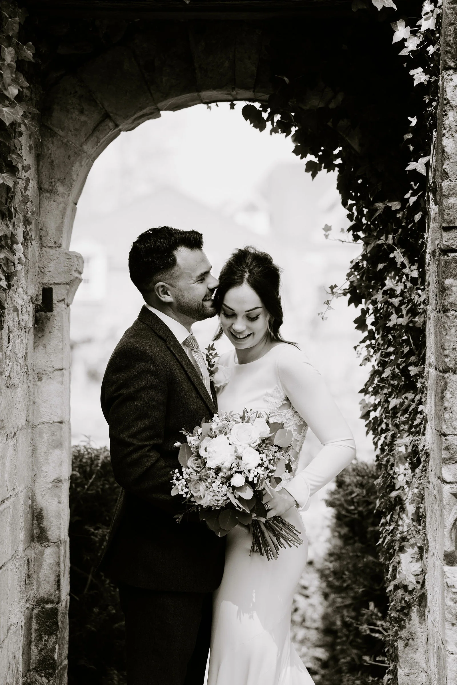 In black and white, a groom and a bride hold each other with her bouquet between them beneath a stone arch hung with ivy at Lainston House
