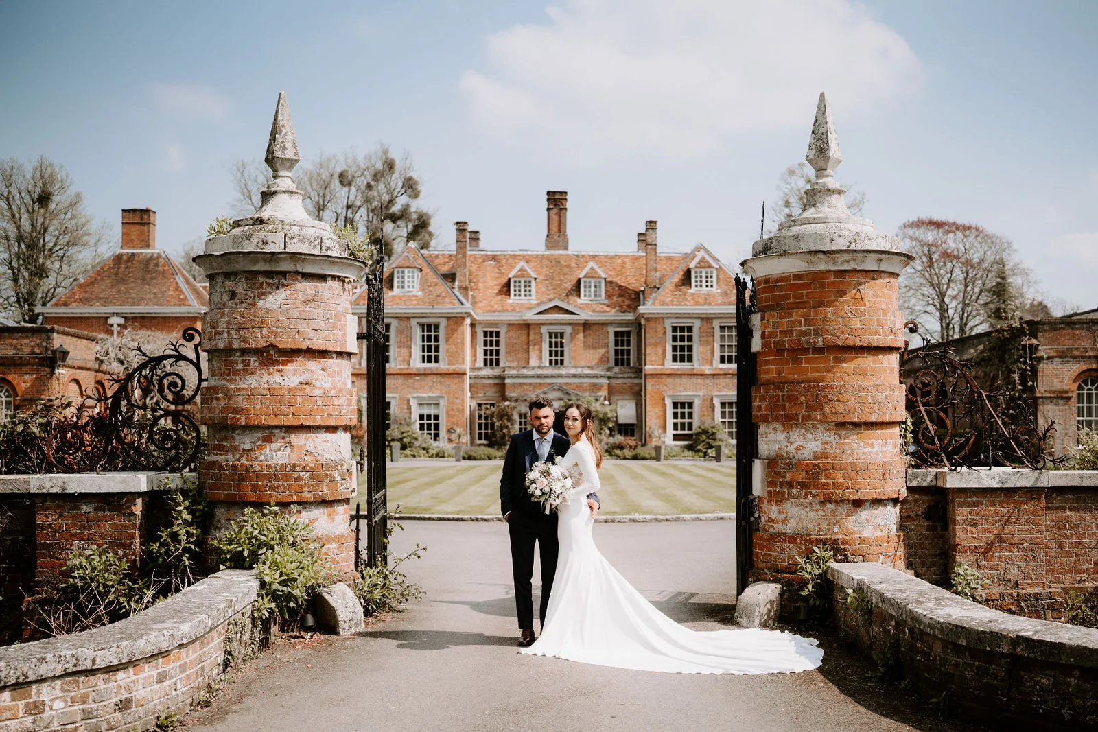 A bride and groom stand between two brick gate piers on the drive with the red-brick house of Lainston House behind them