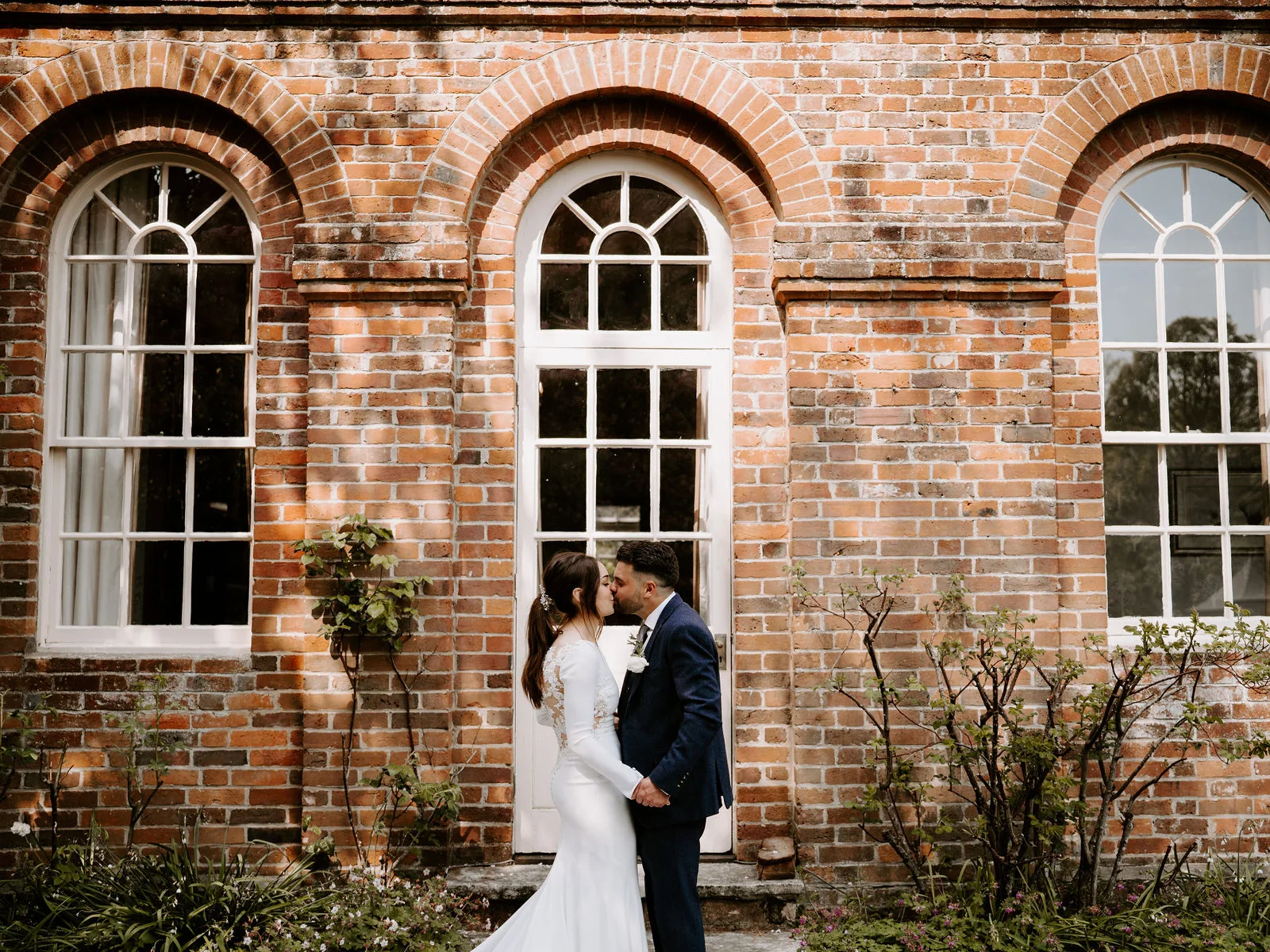 A bride and groom kiss in front of tall white-framed arched windows in a red brick wall at Lainston House