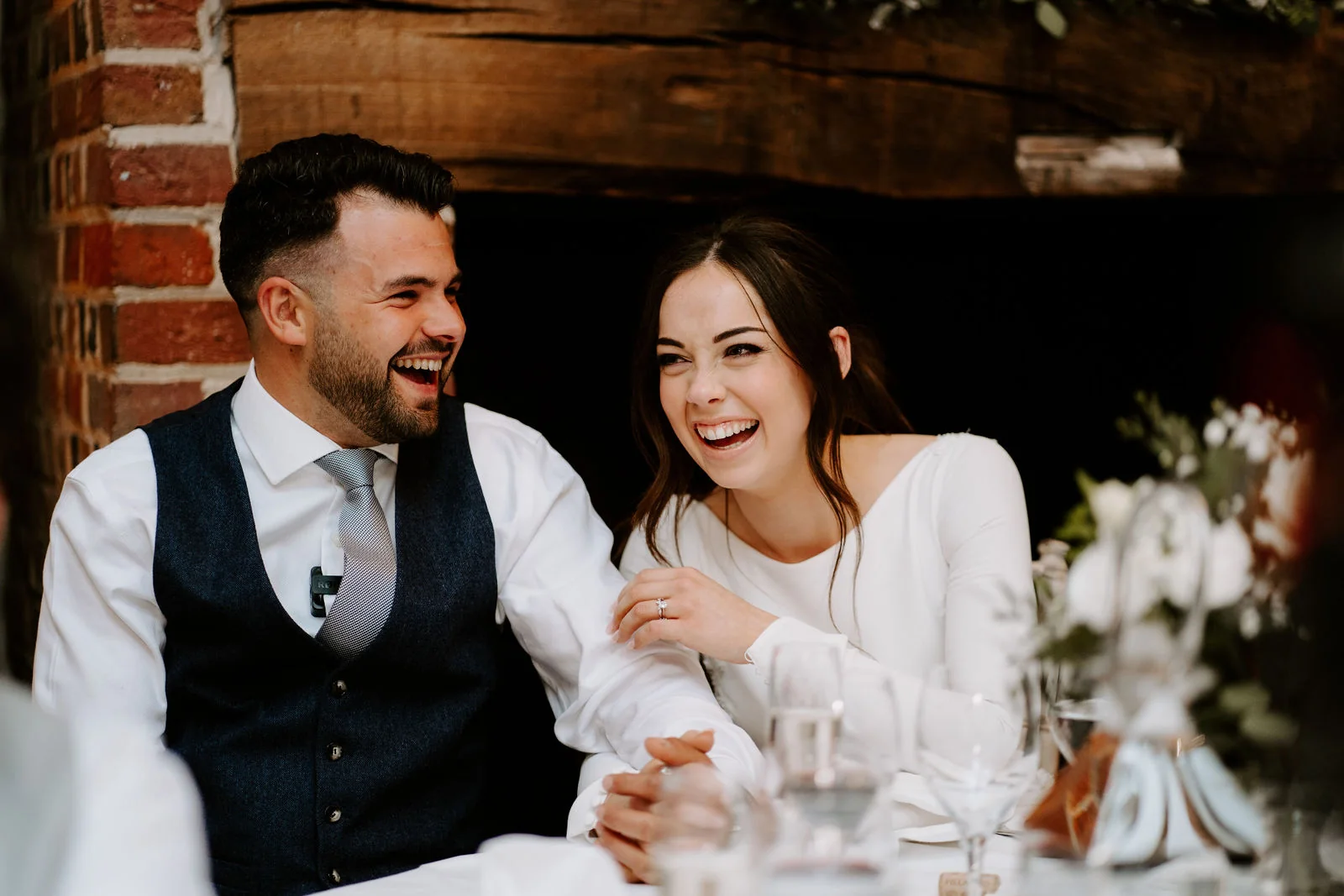 A groom laughs as his bride leans close beside him at a table among flowers in a brick-walled room at Lainston House