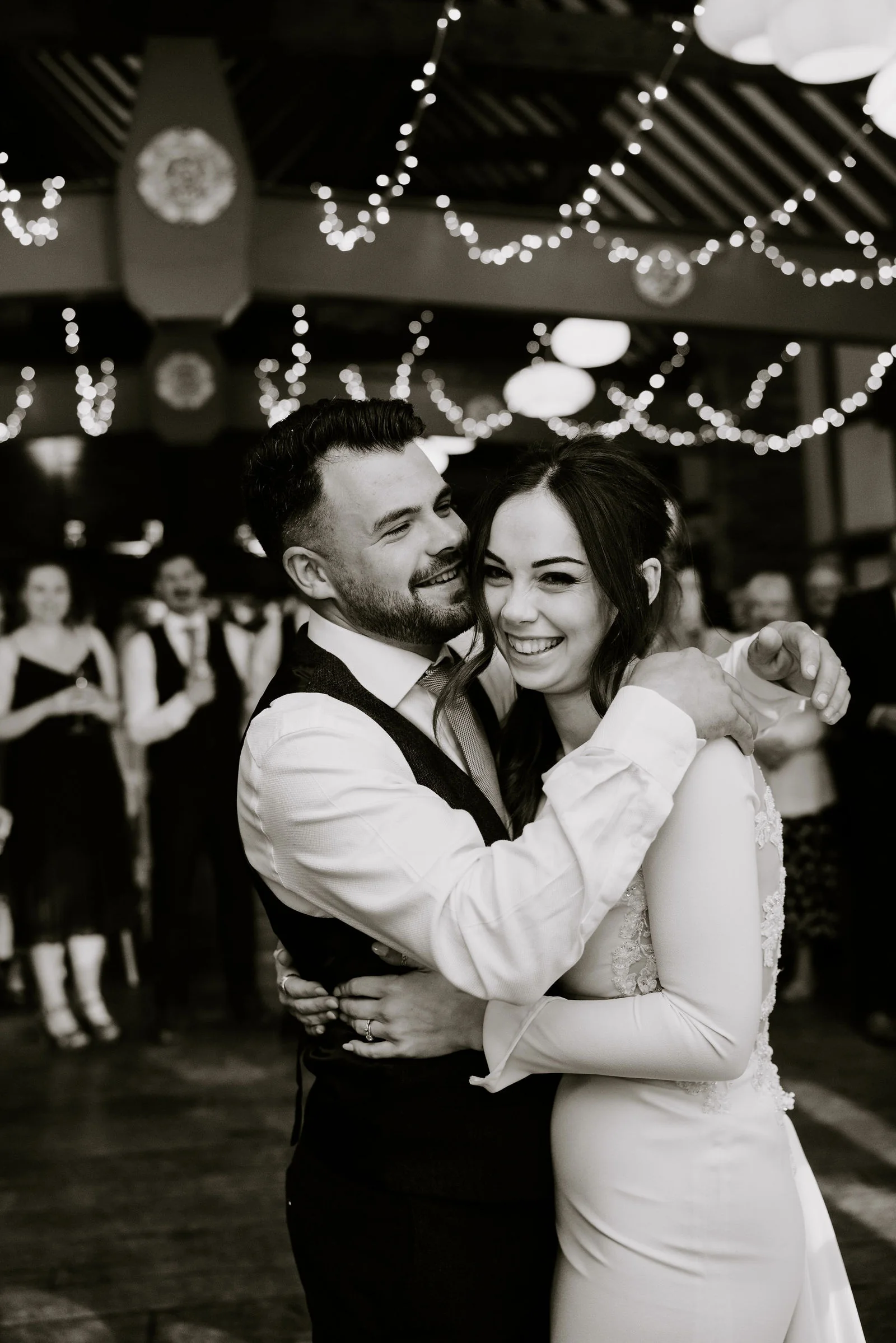 In black and white, a groom and a bride dance close together beneath string lights at Lainston House