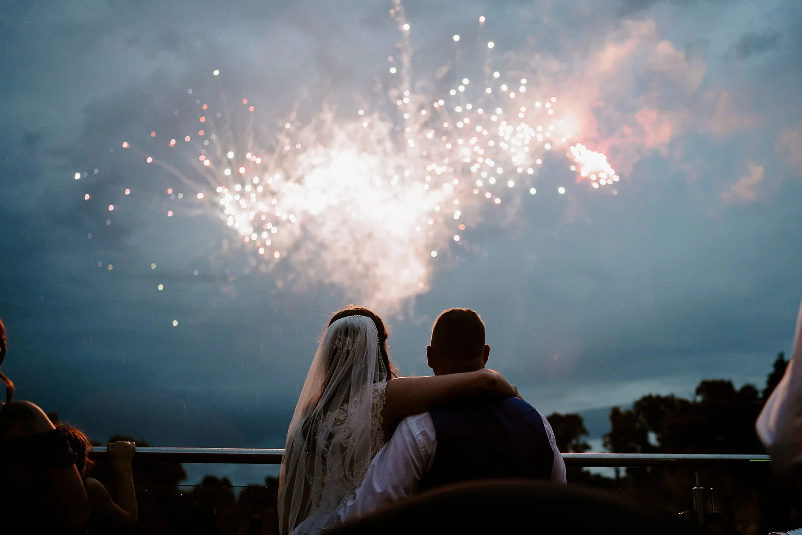 A bride and groom seen from behind with their arms around each other watch fireworks burst in a darkening sky at Old Thorns