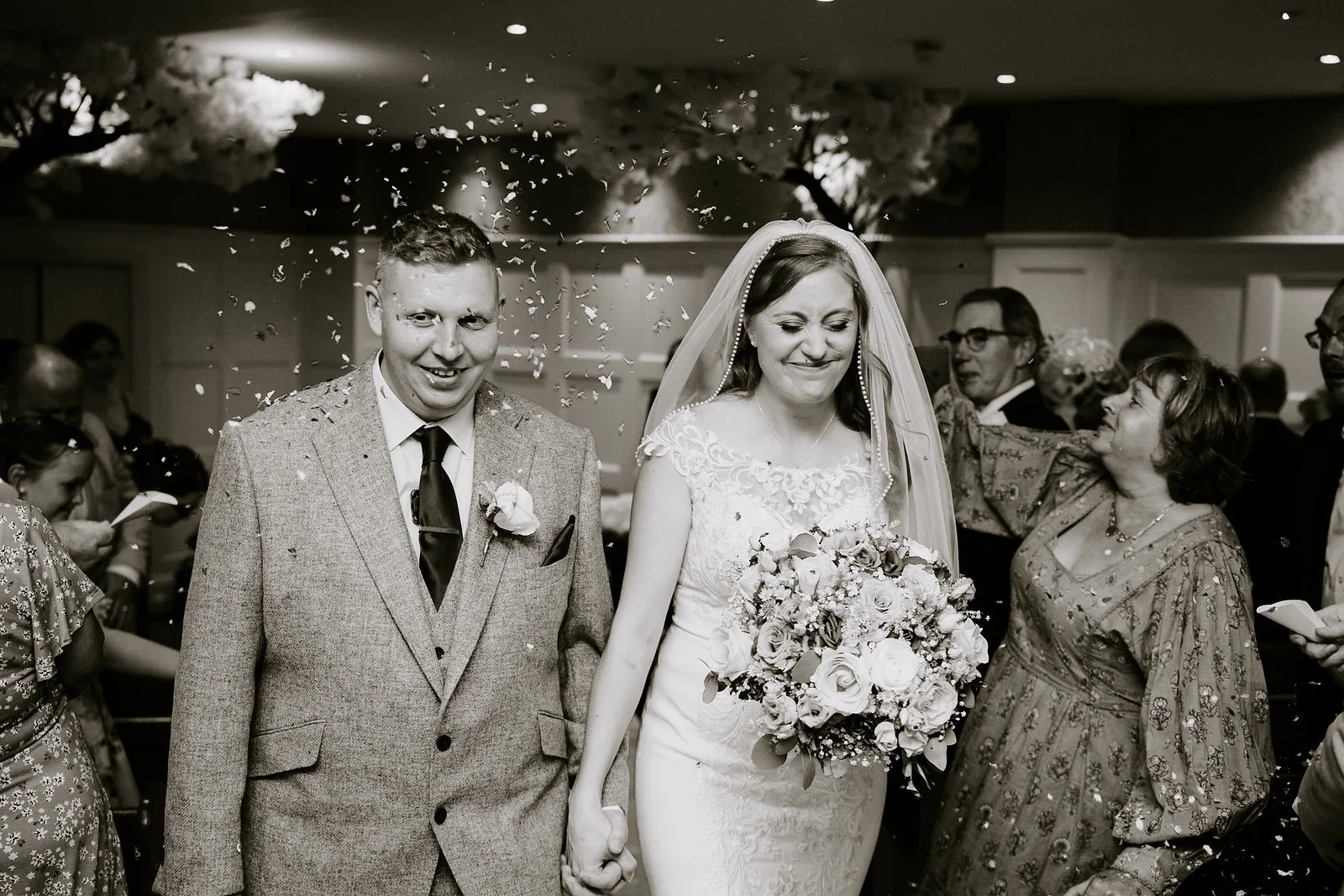 In black and white, a bride with a bouquet and a groom in a tweed suit walk smiling through a crowd of guests at Old Thorns