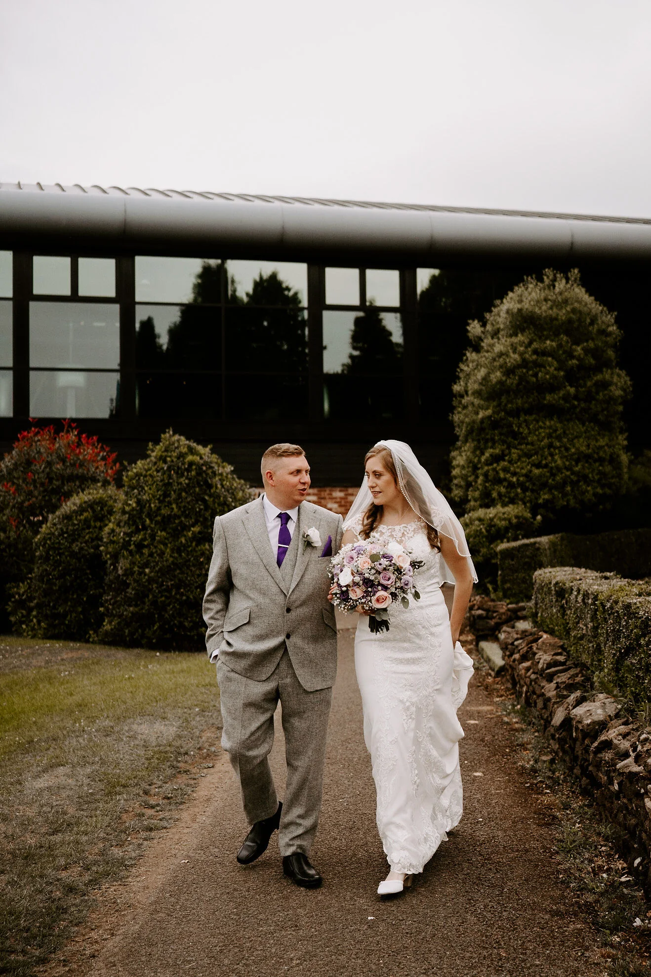 A groom in a grey suit and a bride in a veil walk along a garden path with the hotel building behind them at Old Thorns