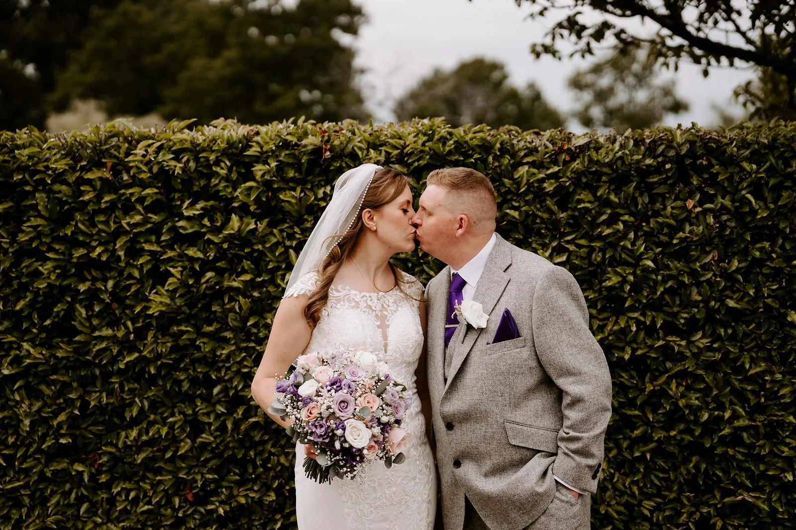 A bride in a veil holding a bouquet kisses her groom in a grey suit in front of a clipped hedge at Old Thorns