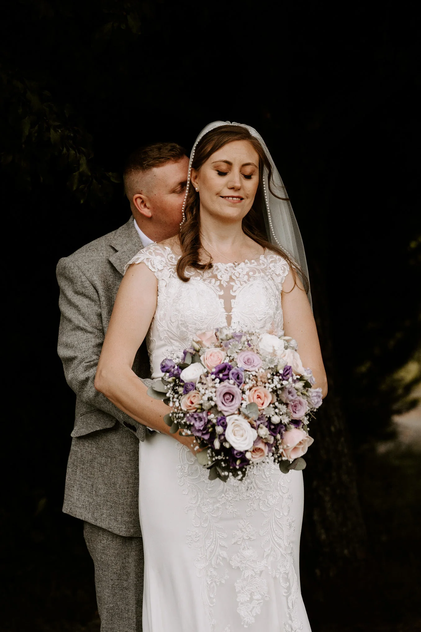 A bride with her eyes closed holds a bouquet of lilac flowers as her groom holds her from behind against dark foliage at Old Thorns