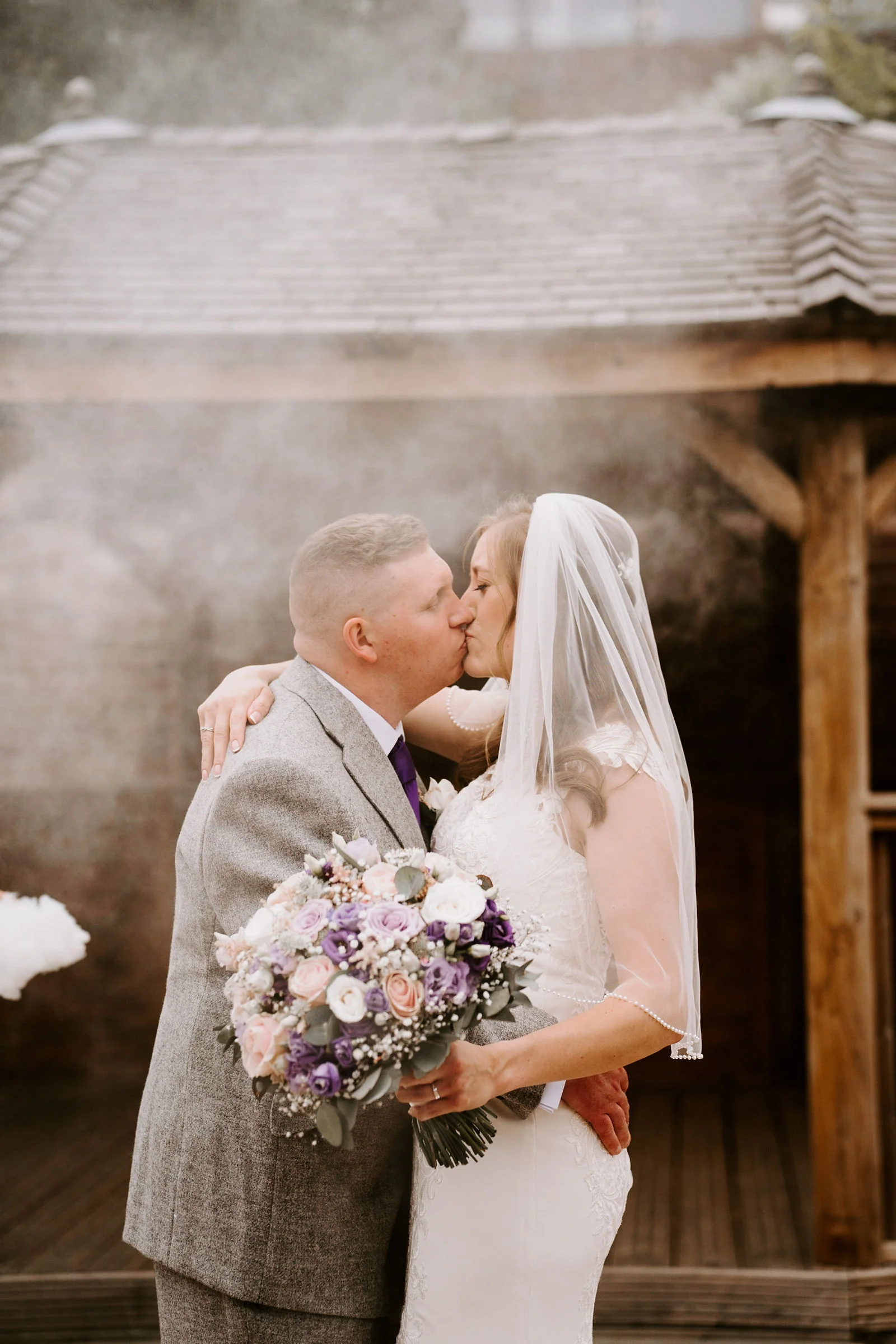A bride holding a bouquet and her groom kiss beside a timber shelter as smoke drifts around them at Old Thorns